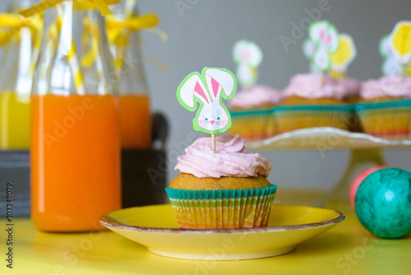 Fototapeta Colorful cupcake decorated with bunny picker on the yellow plate and orange juice in glasses in Easter scene 