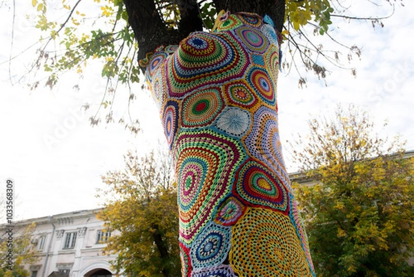 Fototapeta A tree wrapped in colorful crochet yarn art, showcasing vibrant patterns against a backdrop of an urban park. A unique blend of nature and creative street art in autumn