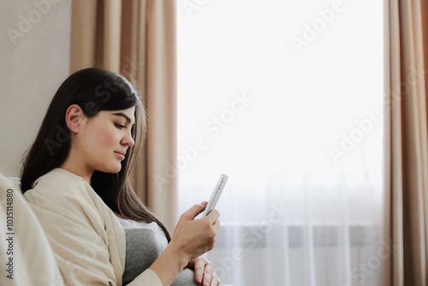 Fototapeta Charming pregnant woman with baby belly touch mobile phone, do online shopping, browse internet, scroll website, watch podcast for future moms, texting, use telemedicine app while sitting at home