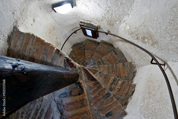 Fototapeta A spiral staircase with worn brick steps and a curved metal railing. The walls are whitewashed, with a small window casting light on the stairs. A support beam is visible on the left side of the image