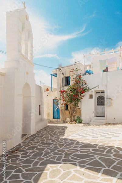 Obraz A tranquil Greek courtyard features beautifully whitewashed buildings and a small church with a bell tower, surrounded by vibrant plants under bright blue skies.