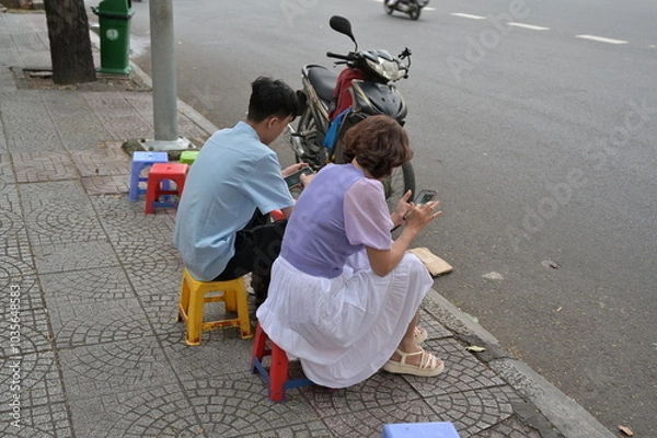 Fototapeta A young couple in love sits on a street in Hanoi-Vietnam while busying themselves with their smartphones.