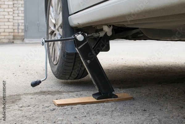 Fototapeta The front wheel of the car is jacked up for replacement. A board is placed under the jack