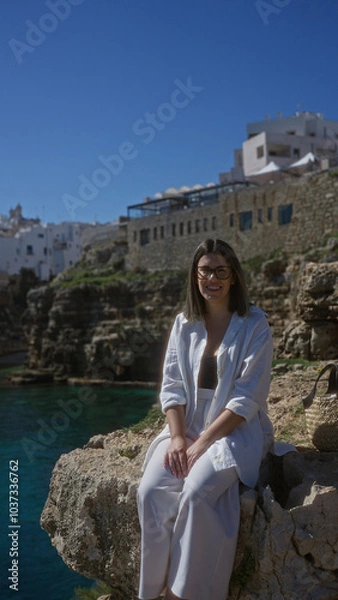 Fototapeta A beautiful young hispanic woman smiling on a rock cliff overlooking the sea at polignano a mare, puglia, italy, with a charming historic beach town in the background.