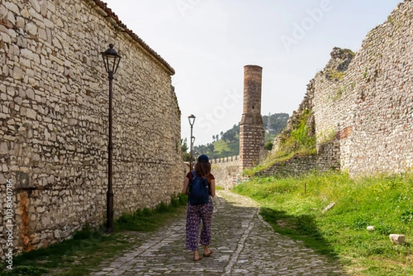 Fototapeta Tourist woman walking in charming cobblestone alleyway towards ancient red mosque in Berat castle, Albania. Weathered stone walls partially collapsed creating sense of history and decay. Xhamia e Kuqe