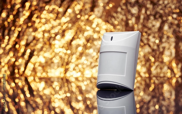 Fototapeta White plastic alarm motion sensor with fancy shiny gold background full of sparks out of focus.
