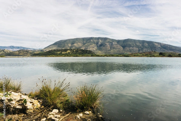 Fototapeta Mountain lake with blue sky. Clear water landscape. Calm lake surface with mountains on the horizon. Beautiful nature background with summer vibes. Holiday in Albania. Vacation trip to Butrint.