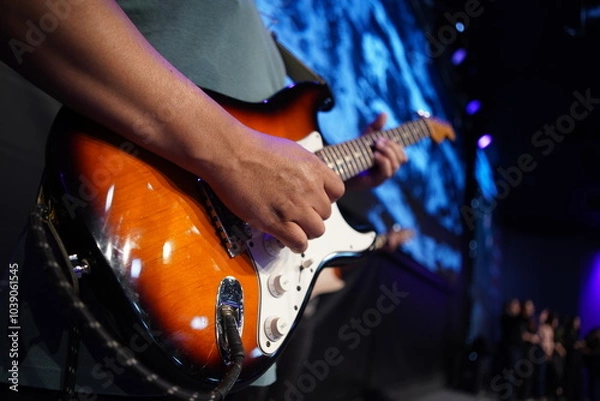 Fototapeta Close-up of a classic redwood Stratocaster guitar, with black edges, held by a White man's arms. The rich texture and warm tones capture the essence of musical passion.
