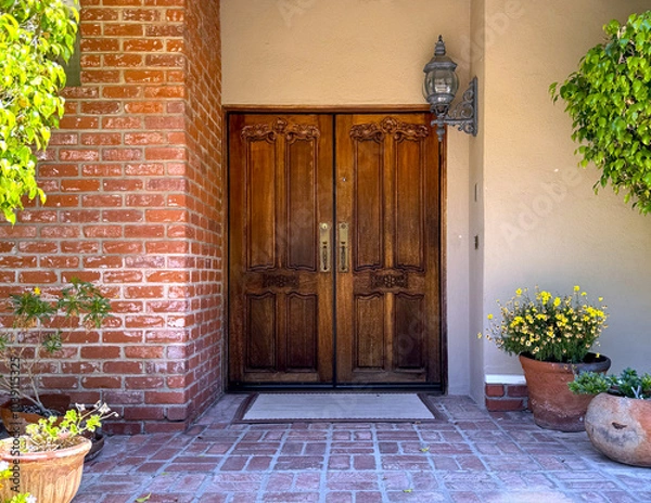 Obraz front door of a American house: wooden double doors with intricate carvings and brass doorknobs; brick wall, stucco, rustic antique lantern, welcome mat, potted flowers and plants and trees