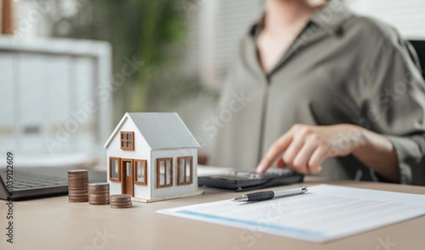 Fototapeta residential, rental, finance, wealth, calculator, agreement, mortgage, property, real, rent. A woman is sitting with calculator and house model. She is pointing at the calculator and the house model.