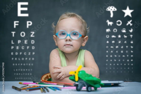 Fototapeta Ребенок сидит за столом на котором разбросаны цветные карандаши, на фоне размытом таблицы для проверки зрения 
