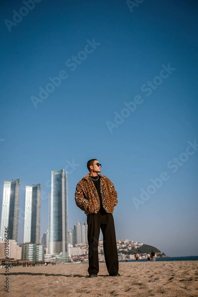 Fototapeta A Korean guy in a leopard fur coat walks along the beach against the backdrop of the autumn landscape of Busan Haeundae.