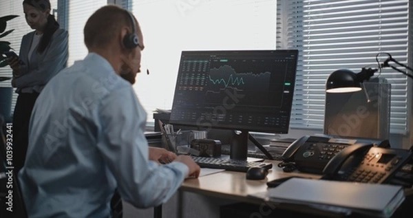 Fototapeta In the stylish modern office: Male financial trader in headset works on computer, talks to business client on call, monitors stocks and share market, takes notes. International stock exchange company.