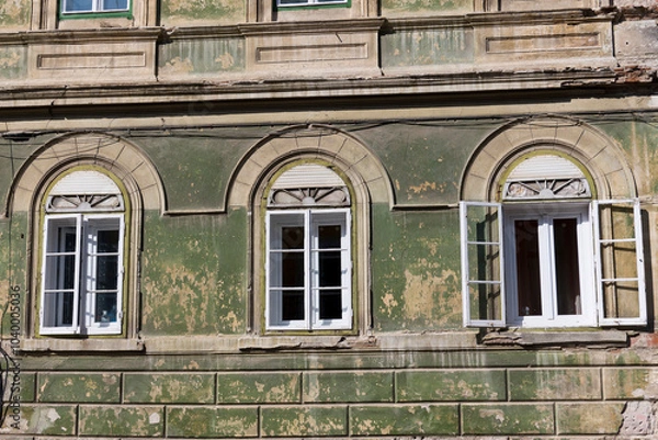 Fototapeta A weathered building facade in Sibiu, Romania, featuring three arched windows with white frames and shutters. The building is painted in a muted green and has peeling paint in several areas. 