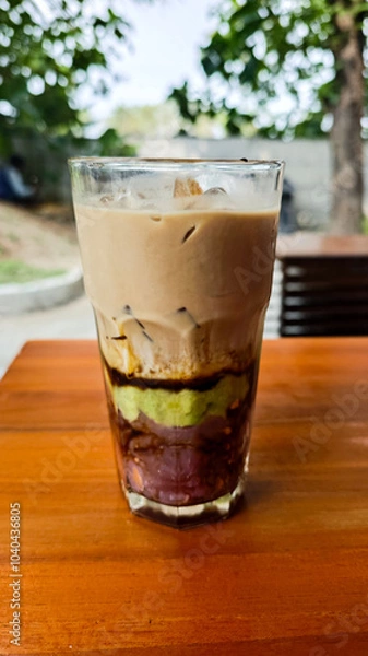 Fototapeta Layered Avocado Mocha Drink with Ube and Sweet Toppings in a Glass on a Wooden Table in an Outdoor Setting