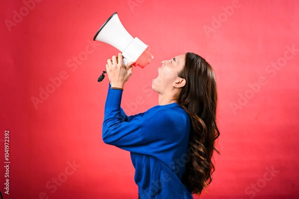 Fototapeta An Asian woman wearing a blue sweater is holding a red and white megaphone while speaking into it. She appears to be shouting or making an announcement. The background is solid red