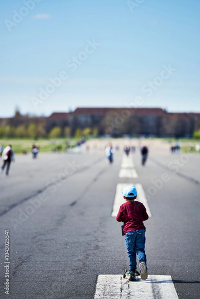 Fototapeta Ready steady go : child riding a scooter on a large airport runway. Kid pretends to take off like an airplane on a airfield tarmac or road conveys a life journey just started. Stamina or speed concept
