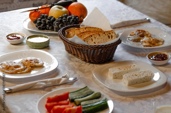 Fototapeta A cozy breakfast spread featuring a mix of fresh fruits, sliced vegetables, bread, cheese, and pancakes. The table is set with a rustic wicker basket for bread, creating a warm atmosphere 