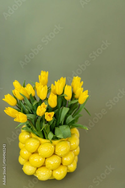 Fototapeta A creative floral arrangement of vibrant yellow tulips styled in a charming lemon-shaped vase against a soft green backdrop