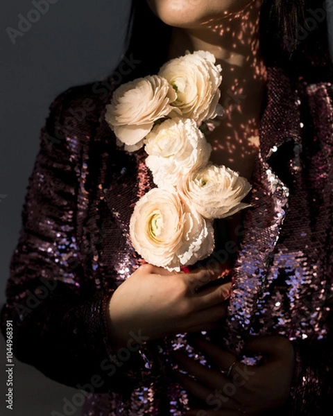 Fototapeta A woman in a shimmering blazer holds delicate white flowers against a backdrop of soft, dappled light, creating a moment of elegance