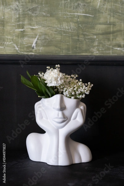 Fototapeta A creative ceramic planter shaped like a face, adorned with delicate white flowers, sitting against a textured green wall