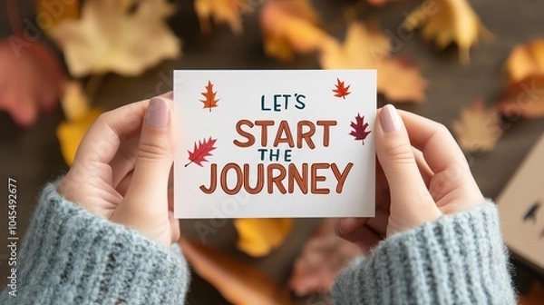 Fototapeta Person holding a card with 'Let's Start the Journey' text surrounded by autumn leaves, symbolizing new beginnings and seasonal change.