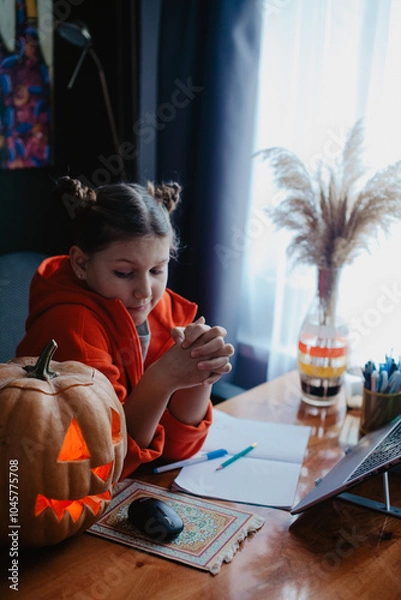 Fototapeta Caucasian happy girl orange color cloths near carved Halloween pumpkin studying online laptop