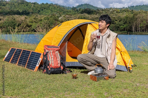 Fototapeta An Asian man sitting and drinking coffee near camping tent beside the lake and mountain in the sunshine morning with backpack and mobile solar cell