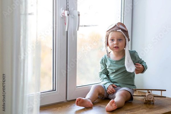 Fototapeta A cute 2-year-old boy in a cap is playing on the windowsill with a wooden airplane. Children's wooden toys for child development.