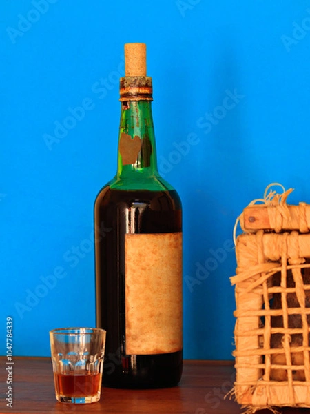 Obraz bottle of a very old wine arranged between a glass with a shot and a wicker basket, on a wooden top, against a blue background.