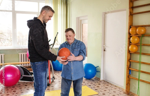 Obraz A man with Down syndrome is working out in a gymnasium with his trainer. They are using a medicine ball