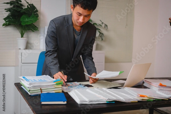 Fototapeta A businessman sits at his desk, working late into the night on his laptop. He reviews documents carefully, showcasing commitment and diligence as he works overtime to meet deadlines
