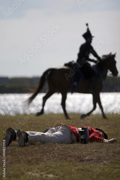Fototapeta Napoleonic era soldier lies dead in a battle re-enactment whilst mounted soldier rides in background.