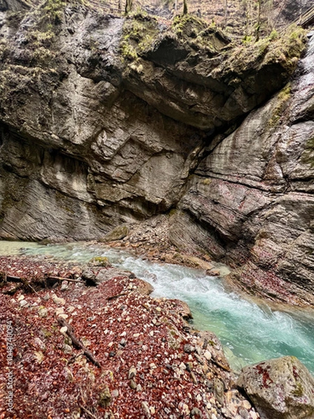 Fototapeta Wet rocks line the vibrant turquoise stream flowing through Partnach Gorge, with red leaves scattered along the ground, creating a striking autumn contrast in the Bavarian Alps.