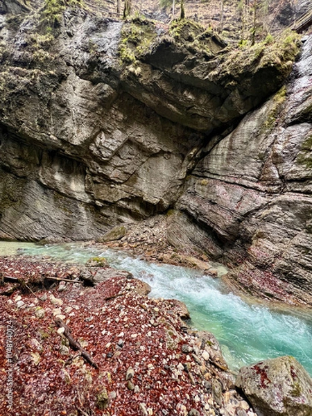 Fototapeta Wet rocks line the vibrant turquoise stream flowing through Partnach Gorge, with red leaves scattered along the ground, creating a striking autumn contrast in the Bavarian Alps.