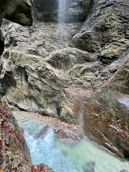 Fototapeta A path winds through the rocky cliffs of Partnachklamm Gorge, with a stream at its base and misty droplets from nearby waterfalls creating an enchanting atmosphere, Garmisch-Partenkirchen.