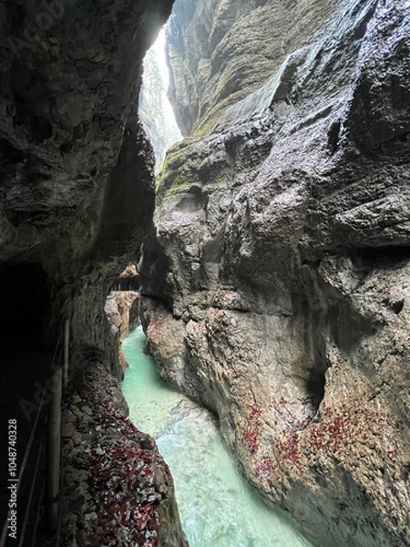 Fototapeta A narrow walkway winds through the rock cliffs at the valley's base alongside a flowing stream at Partnachklamm Gorge, Garmisch-Partenkirchen, offering a stunning alpine experience.