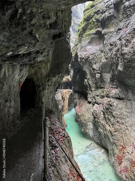 Fototapeta A narrow walkway winds through the rock cliffs at the valley's base alongside a flowing stream at Partnachklamm Gorge, Garmisch-Partenkirchen, offering a stunning alpine experience.