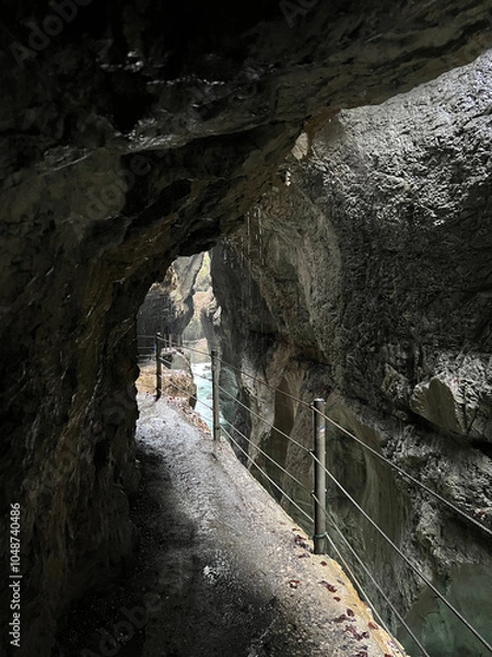 Fototapeta A narrow walkway winds through the rock cliffs at the valley's base alongside a flowing stream at Partnachklamm Gorge, Garmisch-Partenkirchen, offering a stunning alpine experience.