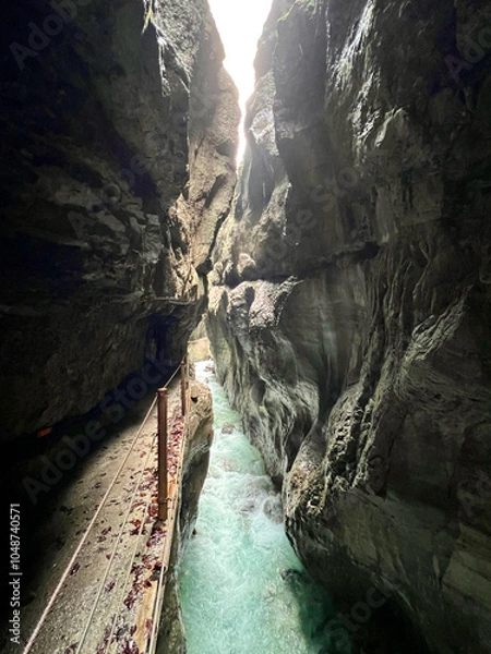 Fototapeta A narrow walkway winds through Partnachklamm Gorge alongside the flowing stream at the valley's base, nestled between dramatic cliffs and lush greenery in Garmisch-Partenkirchen.