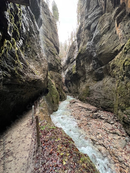 Fototapeta A scenic pathway follows the stream as it flows through the deep valley of Partnachklamm Gorge, surrounded by rocky cliffs and lush greenery in Garmisch-Partenkirchen, Germany.