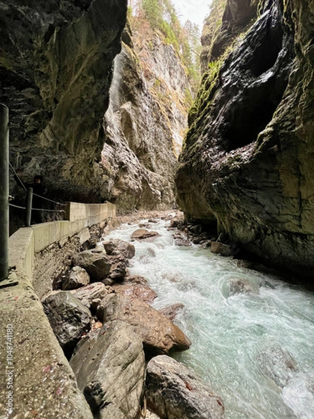 Fototapeta A narrow walkway winds through the rock cliffs at the valley's base alongside a flowing stream at Partnachklamm Gorge, Garmisch-Partenkirchen, offering a stunning alpine experience.