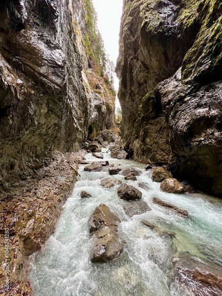 Fototapeta A powerful stream flows through the rocky riverbed of Partnachklamm Gorge, winding between towering cliffs and creating a dramatic, rugged valley scene in the Bavarian Alps.