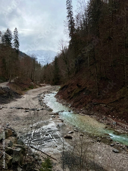 Fototapeta A stream flows from the valley of Partnachklamm Gorge into a lush forest with the majestic mountain range as a backdrop, creating a serene, natural scene in Garmisch-Partenkirchen.