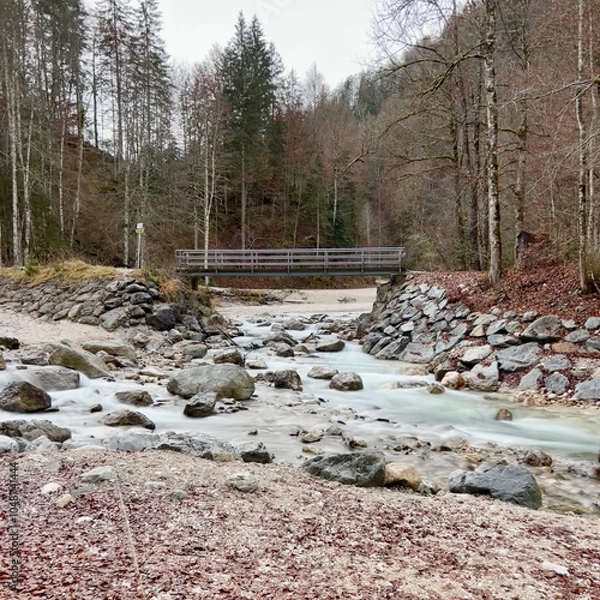 Fototapeta A small bridge near Partnacher Felsentürme spans a clear river, framed by a misty forest and towering mountains, creating a serene, mystical scene at Partnachklamm Gorge.