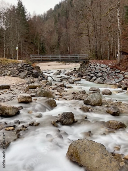 Fototapeta A small bridge near Partnacher Felsentürme spans a clear river, framed by a misty forest and towering mountains, creating a serene, mystical scene at Partnachklamm Gorge.