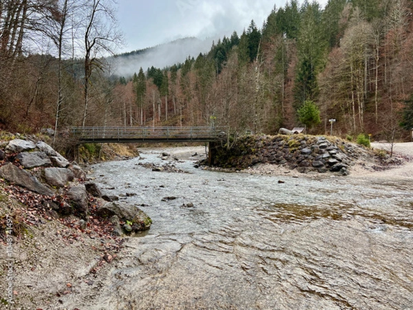 Fototapeta A small bridge near Partnacher Felsentürme spans a clear river, framed by a misty forest and towering mountains, creating a serene, mystical scene at Partnachklamm Gorge.