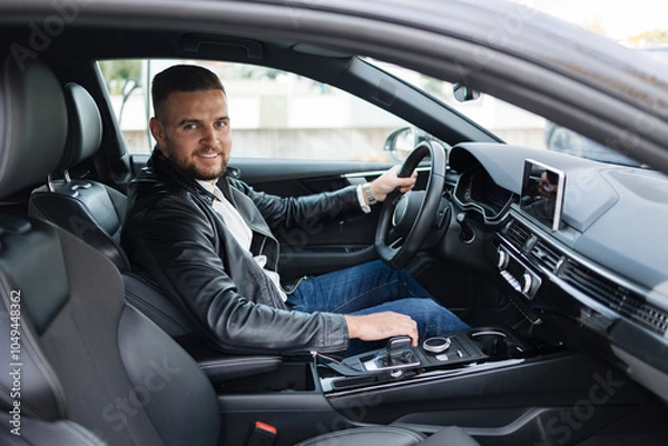 Fototapeta Success in motion. Handsome young man in leather jacket smiling while driving a car. Side view from inside