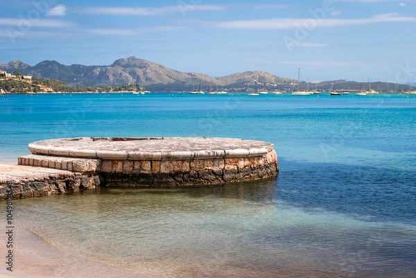 Fototapeta Port de Pollença beachfront in Mallorca with a circular stone pier extending into the calm, shallow waters of the bay, framed by distant Serra de Tramuntana mountains, for tourism and travel content.