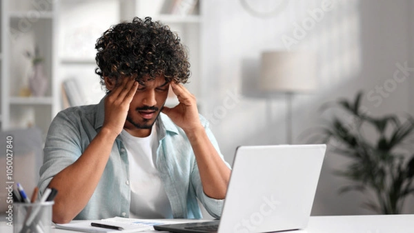 Obraz A young Indian male businessman is sitting at a desk trying to work, checking schedules and getting frustrated that something is not working out. Headache, fatigue, burnout.